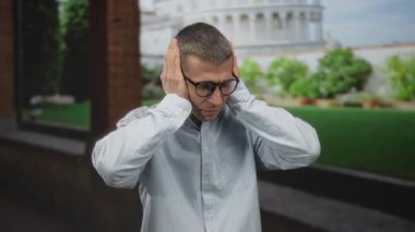 Dışarda dikilen bir adam şok olmuş bir ifadeyle, gözlüklü ve çizgili gömlekli, gündüz vakti resim gibi bir italyan sokak ortamında.