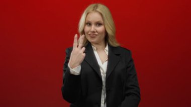 Siyah ceketli bir kadın kırmızı arka plana vurmaya karşı hashtag jesti yapıyor, sosyal medyada kendine güvenen ifade ve sarı saçla iletişim kuruyor.