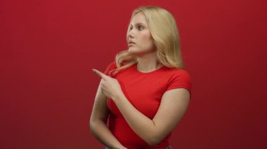 Kırmızı tişörtlü, sarı saçlı genç bir kadın canlı kırmızı bir arka plana karşı jest yapıyor, yön ve karar veriyor..