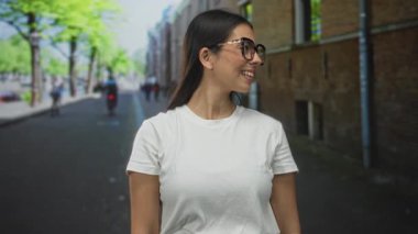 Büyük boy kaplumbağa kabuğu gözlüklü ve beyaz tişörtlü bir kadın, bir şehir caddesinde tuğla bir binanın yanında dururken gülümsüyor ve başparmağını kaldırıyor..