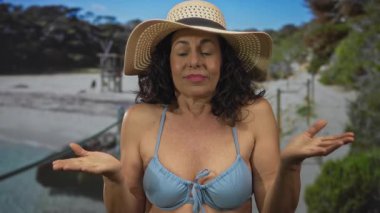 Mavi bikinili ve şapkalı orta yaşlı İspanyol bir kadın güneşli sahilde, arka planda doğal yeşillik ve deniz kenarında rahatlık yayıyor..