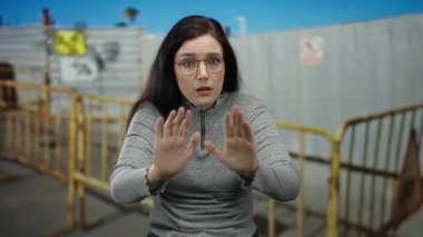 Gözlüklü bir kadın inşaat alanında duruyor. Metal bariyerlere ve mavi gökyüzüne karşı. Gri kazak ve kahverengi saçlar giyiyor..