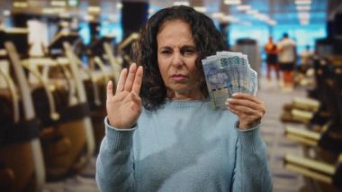 Orta yaşlı bir kadın, spor salonunda Güney Afrika Rand banknotları tutuyor. Egzersiz ekipmanları ve hareketli bir geçmişi olan, kapalı alandaki şüphelerini dile getiriyor..