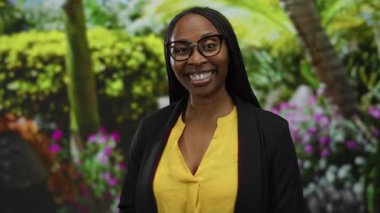 Canlı bir parkta gülümseyen bir kadın, çiçekler takan, siyah ceketin altında sarı bir bluz giyen, açık havada neşeli bir sahne sergiliyor..