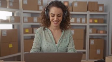 Ofis çalışma alanında istiflenmiş paket kutularının arasında kulaklık takan ve gülümseyen ve dizüstü bilgisayarda yazan kadın; üretkenlik küçük işletme ekommersi.