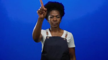 Önlük giymiş, mavi arkaplanı işaret eden, odaklanmış bir ifade veren, stüdyo ortamında izole edilmiş bir kadın..