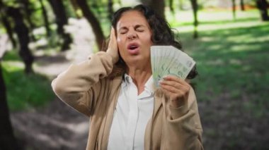 Orta yaşlı bir kadın parkta bir dolar tutuyor, kulağını bir rahatsızlık ifadesi ile kapatıyor, yeşillik ve ağaçlarla çevrili..