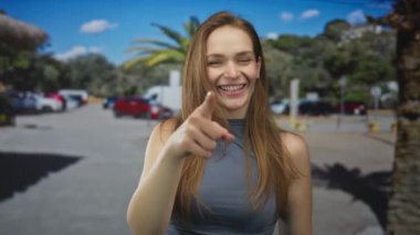 Gülümseyen yüzü olan beyaz bir kadın, şehrin otoparkında kamerayı işaret ediyor..