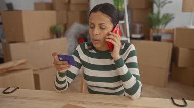 Evin içinde taşınan kutularla çevrili telefon ve kredi kartı kullanan bir kadın, iletişim ve çoklu görev yaparken yeni bir ev veya apartman dairesine taşındığını öne sürüyor..