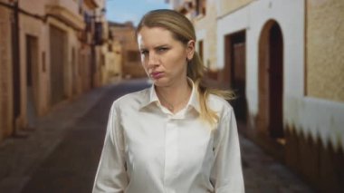 Beyaz gömlekli bir kadın, tarihi bir sokak ortamında hoşnutsuz bir ifade sergiliyor, kırsal bir mimarinin arka planına karşı duygusal bir an yakalıyor..