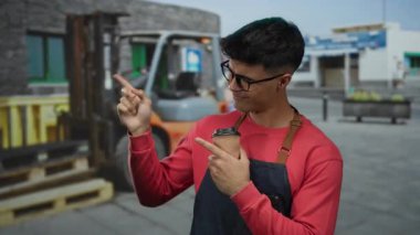 Elinde kahve fincanı olan genç bir adam forklift arka planında açık havada gülümsüyor, kırmızı kazak ve önlük giyiyor..