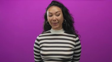 Çizgili bluzlu bir kadın mor stüdyoda elini ve dilini kullanarak yüzüne dokunuyor..