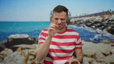 Kulaklık takan ve çizgili gömlek giyen genç adam neşeli bir şekilde güneşli bir sahilde jestler yapıyor. Arka planda mavi deniz ve kayalık bir kıyı var.