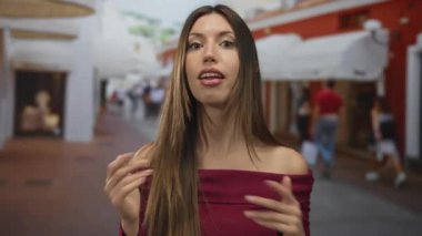 Genç bir kadın, yoğun bir şehir caddesinde dükkanların önünde el kol hareketi yaparken omuz üstü açık bir bordo ile çıplak omzunu gösteriyor..