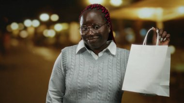 Gece şehir sokağında elinde alışveriş çantası olan bir kadın arka planda ışıklarla açık şehir manzarasını neşeli bir ifade ve moda tarzıyla gösteriyor..