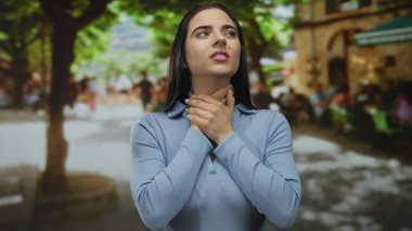 Mavi tişörtlü genç bir İspanyol kadın. Yoğun bir sokakta, arka planda bulanık yeşillik ve insanlarla boğazını sıkıyor..