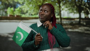 Pakistan bayrağını parkta elinde tutan kadın, arka planda ağaçlarla düşünceli bir ifade ve parlak gün ışığında rengarenk saç stili ile kültürel çeşitliliği öne sürüyor..