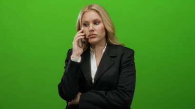 Akıllı telefondan konuşan bir kadın yeşil bir arka plana karşı takım elbise giyiyor, odaklanmış ve sohbete dalmış görünüyor, profesyonel iletişim ve modern iş kıyafetleri sergiliyor..