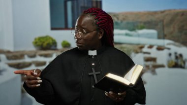 Gözlüklü ve papaz kıyafeti giyen kırmızı örgülü bir kadın kitabı ve puanları elinde tutarken açık havada, yeşil ve taş basamaklı binaların yanında duruyor..