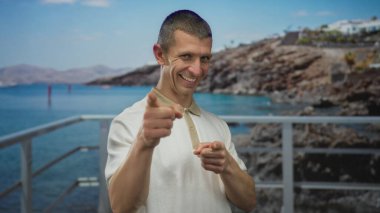 Güneşli bir sahilde gülümseyen bir adam kayalık tepeler ve açık denizle mavi gökyüzünün altında.