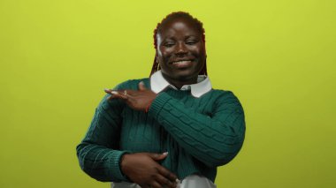 İzole edilmiş sarı bir arka plana gülümseyen, yeşil bir kazak giyen, hareketleriyle kendine güvenini ve güvenini ifade eden bir kadın..