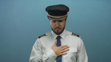 Sakallı ve üniformalı genç pilot, kafa karışıklığını ifade eden, havayolu, İspanyol ve profesyonel havacılık temalarını vurgulayan mavi bir arka plana karşı duruyor..