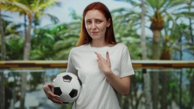 Parkta kızıl saçlı bir kadın elinde bir futbol topu tutuyor, kafası karışmış ve beyaz bir gömlek giyiyor, canlı yeşillikle çevrili..