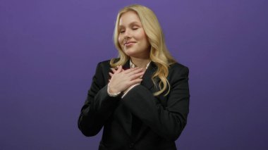 Minnettarlığını ifade eden bir kadın, elleri göğsünde koyu mor bir arkaplan karşısında, resmi siyah bir spor ceketle, samimi bir duyguyu somutlaştırıyor..