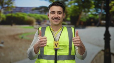 Yansıtıcı yelek giyen İspanyol adam güneşli bir park ortamında gönüllü ruhu ve açık hava toplum nişanını sembolize ediyor..