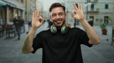 Sakallı ve kulaklıklı genç adam şehrin arka planında bulanık bir jest yaparken gülümsüyor. Açık havada tasasız ve rahat bir atmosfer izlenimi veriyor..