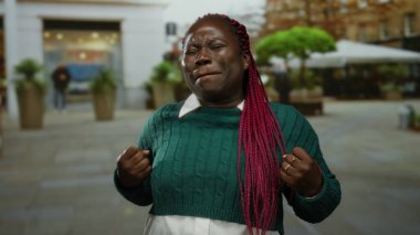 Kalabalık bir caddede yeşil bir kazakla gülümseyen kadın, arka planda canlı renkler ve hareketli bir atmosferi olan şehir ortamında heyecan gösteriyor..