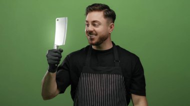 Önlük takan sakallı genç adam eldivenli bir baltayı yeşil bir duvara doğru tutuyor ve sırıtıyor..