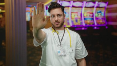 Sakallı ve steteskoplu genç beyaz erkek doktor kumarhanede duruyor. Arka planda kumar makineleri var. Sağlık ve kumar ortamlarının benzersiz bir zıtlığını oluşturuyor..