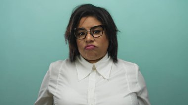 Gözlüklü ve beyaz gömlekli kadın, modern tarz ve duygulara odaklanmış saf mavi bir arkaplan karşısında düşünceli bir ifade sergiliyor..