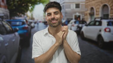 Öğleden sonra güneşli şehir caddesinde ellerini nazikçe göğsüne yaklaştıran İspanyol genç adam..