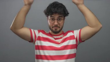 Sakallı ve gözlüklü İspanyol genç adam beyaz arka plana karşı şakacı kulaklar yapıyor, kırmızı çizgili gömlek giyiyor, neşe ve mizah taşıyor..