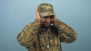 Adam kulaklarını parlak mavi bir stüdyoda kamuflaj asker üniforması ve künye takarak kulaklarıyla kapatıyor..