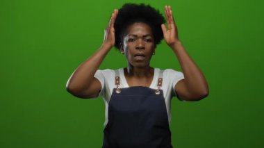 Önlüklü kadın, elleri başının üstünde yeşil arka plana karşı duruyor. Stres ve şaşkınlık taşıyor, beyaz gömlek ve küpeler takıyor..