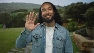 Ormanda el sallayan adam kameraya gülümserken kot ceket ve beyaz tişört, uzun kıvırcık saç ve sakal giyiyor..