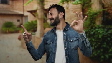 Sakallı İspanyol bir adam yeşil ağaçlarla çevrili güneşli bir parkta jest yapıyor. Kot gömlek giyiyor ve açık havada pozitifliğini gösteriyor..