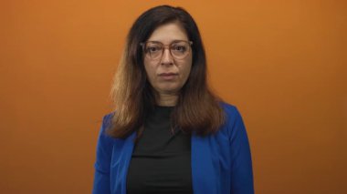 Mavi ceketli İspanyol kadın gözlük takıyor. Stüdyoda parmak göğse turuncu zemin gösteriyor. Sürpriz..