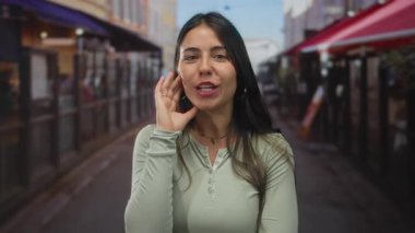 Şehir caddesinde gülümseyen, yeşil elbiseli, uzun siyah saçlı ve rahat duruşu olan bir kadın..