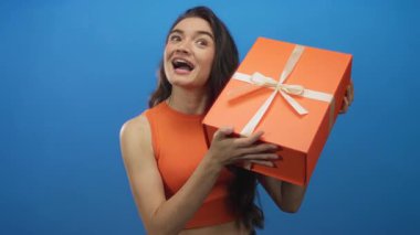 Stüdyoda beyaz kurdeleyle sarılı parlak turuncu hediye kutusu tutan gülümseyen kadın; sevinç kutlaması sürprizi.