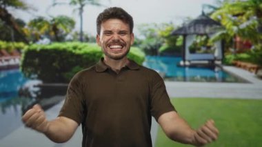 İspanyol asıllı genç adam dişlerini göstererek gülümsüyor ve havuzda sağ kolunu esnetiyor kahverengi polo tişört giyiyor..