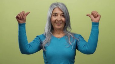 Mavi kazaklı, gri saçlı yaşlı kadın sarı arka planda kendine doğru gülümsüyor..