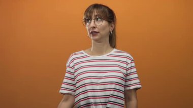 Stüdyoda turuncu duvarlı, çizgili gömlek, yuvarlak gözlük ve halka küpeler takan, kollarını çapraz tutan, şüphecilik meraklısı bir kadın..