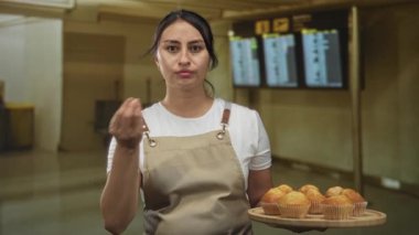 Bir tepsi kek tutan önlüklü kadın havaalanında parmaklarını çimdikleyerek jest yapıyor; neşeli misafirperverlik..