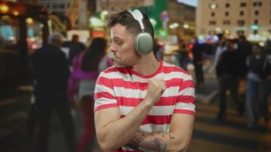 Kulaklıklı genç adam, arka planı bulanık, kırmızı çizgili tişörtlü, ilginç bir ifadeyle başparmağının arkasını gösteren kalabalık bir şehir caddesinde duruyor..