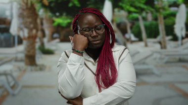 Bir kadın şehir caddesinde elleri çenesinde dikiliyor düşünceli görünüyor, gözlükler takıyor ve örgü örüyor, ağaçlar ve şehir manzarasıyla çevrili..