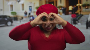 Kırmızı elbiseli bir kadın şehir caddesinde gülümsüyor elleriyle bir kalp oluşturuyor, canlı bir atmosferi olan şehir ortamında mutluluğu sergiliyor..